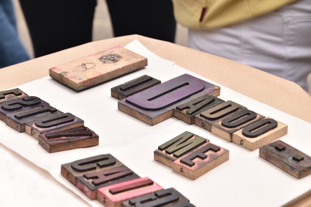 Primer plano de tipos móviles de madera entintados con colores púrpura, negro y verde sobre una mesa de trabajo.